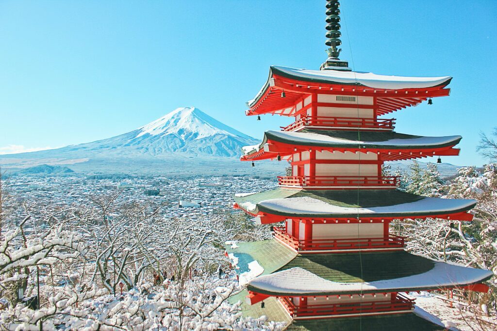 Fujiyoshida Chureito Pagoda surrounded by trees, covered with snow, overlooking the city with Mt. Fuji in the background.